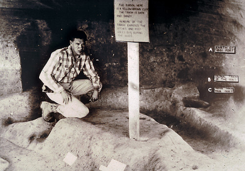 Black-and-white photo of a man kneeling in a trench next to a sign. Markers on the wall behind him indicate layers in the soil.