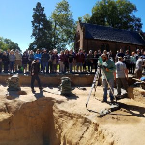 Large group listens to tour guide while archaeologists record finds