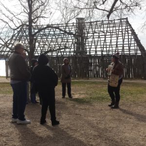 Guide shows photograph to visitors standing in front of reconstructed barracks