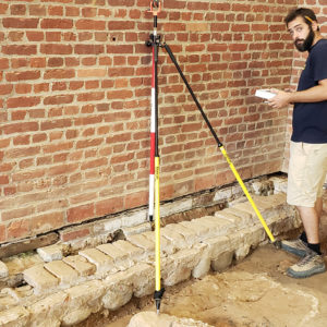 Man stands next to tripod in excavated floor by brick walls