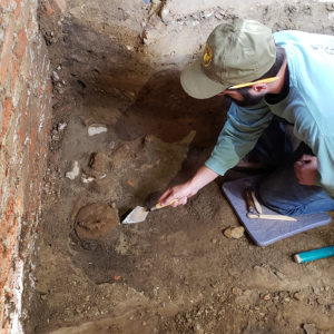 Kneeling archaeologist excavates dirt floor in front of brick wall