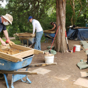 three archaeologists screening for artifacts over wheelbarrows next to a line of trees