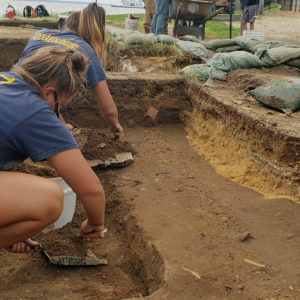 two archaeologists trowel in a unit while others screen for artifacts in the background