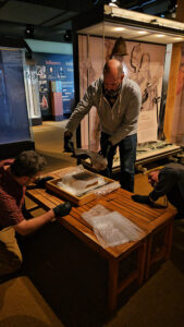Senior Conservator Chris Wilkins, Director of Conservation and Collections Michael Lavin, and Senior Conservator Dan Gamble (L-R) remove the breastplate from the Archaearium exhibit. The breastplate will be treated to remove corrosion and prevent future oxidation.