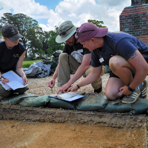 Staff Archaeologist Caitlin Delmas assists field school student Richard Fallon with using a Munsell color chart to classify a soil sample as Archaeological Intern Katherine Griffith records the results.