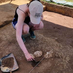 Field school student Sofia Zate bisects a feature in the westmost excavation square in front of the Archaearium. Note the three Chesapecten jeffersonius fossils in situ.