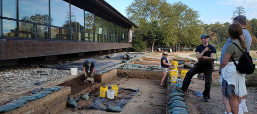 Staff Archaeologist Natalie Reid explains the Archaearium excavations to some visitors. Archaeological Field Technicians Eleanor Robb and Hannah Barch are at work behind her.