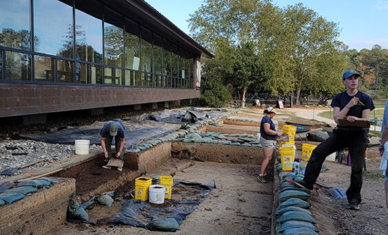 Staff Archaeologist Natalie Reid explains the Archaearium excavations to some visitors. Archaeological Field Technicians Eleanor Robb and Hannah Barch are at work behind her.