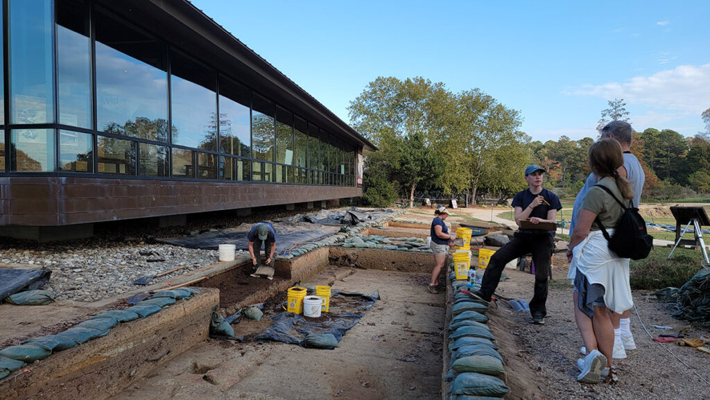 Staff Archaeologist Natalie Reid explains the Archaearium excavations to some visitors. Archaeological Field Technicians Eleanor Robb and Hannah Barch are at work behind her.