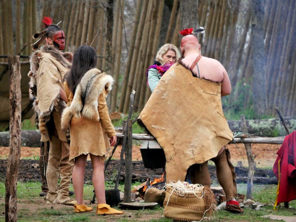 Virginia Indian interpreters wearing historic clothing made of animal hide stand around a fire at Henricus Historical Park