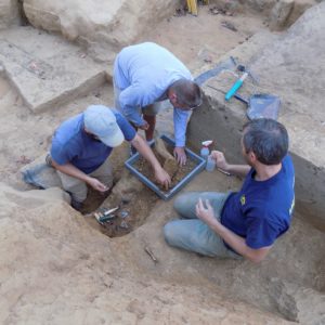 Archaeologists lifting up a tray containing an excavated artifact from a unit