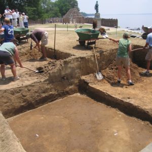 Students shovel dirt from excavation units into wheelbarrows while a tour guide gestures to a small group in the background