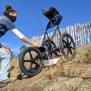 woman steadying a ground-penetrating radar unit as another woman uses a rope to pull it up a low wall