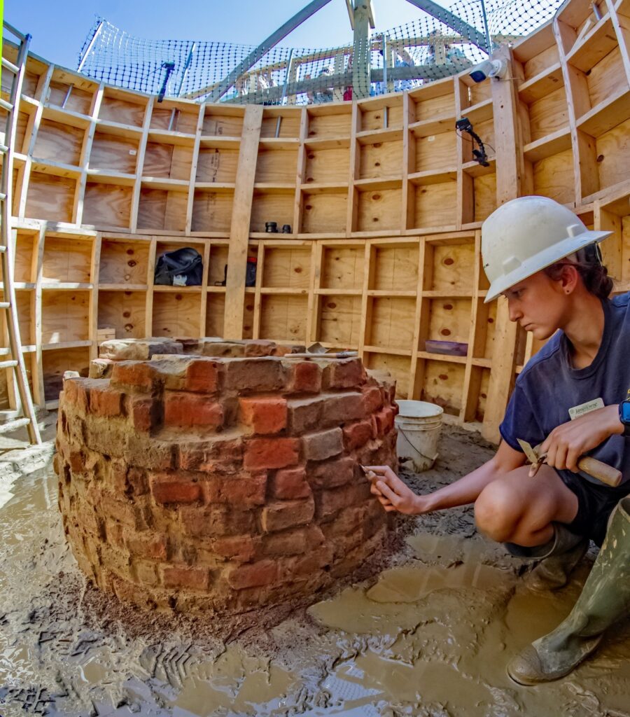 A person in muck boots and a hard hat uses a trowel to gently clean the remaining eight courses of bricks above the excavated ground surface. They are surrounded by a wooden safety frame and the ground surface is very muddy.