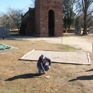 Archaeologist kneeling and pointing to a wooden post with a brick church in the background