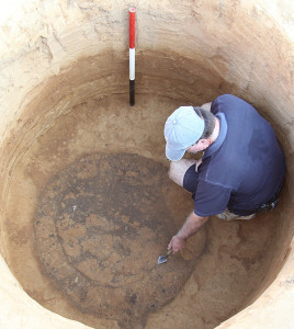 Archaeologist crouching in an excavated well and outlining a feature with a trowel at the bottom