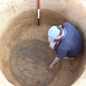 Archaeologist crouching in an excavated well and outlining a feature with a trowel at the bottom
