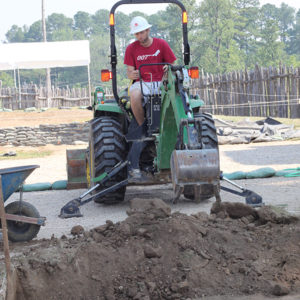 Archaeologist operating a backhoe behind an excavation unit