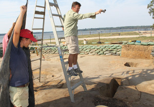 Archaeologist stands on a ladder and holds a camera above an excavation unit as another archaeologist lifts up a tarp to cover the feature with shade