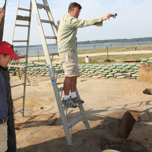 Archaeologist stands on a ladder and holds a camera above an excavation unit as another archaeologist lifts up a tarp to cover the feature with shade