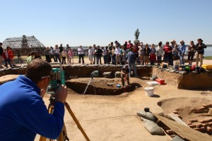 Archaeologist maps feature with a transit while another gives a tour to a crowd in the background
