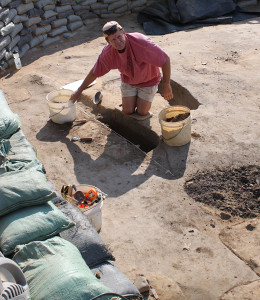 Archaeologist kneeling in an excavation unit and reaching for a bucket of dirt