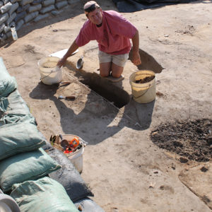 Archaeologist kneeling in an excavation unit and reaching for a bucket of dirt