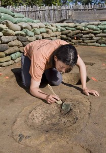 archaeologist brushes off a buried copper square in an excavation area lined with sandbags