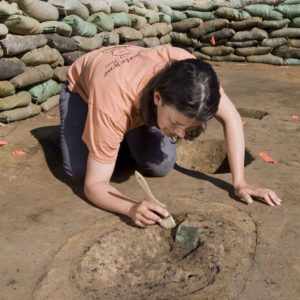 archaeologist brushes off a buried copper square in an excavation area lined with sandbags