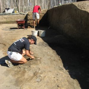 Archaeologist troweling in an excavation unit while another shovels in the background