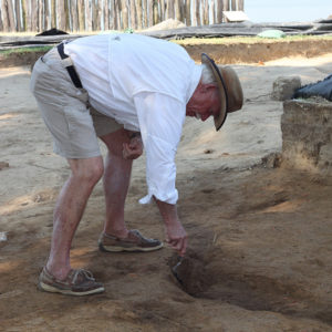 Archaeologist bending over to excavate a feature with a trowel