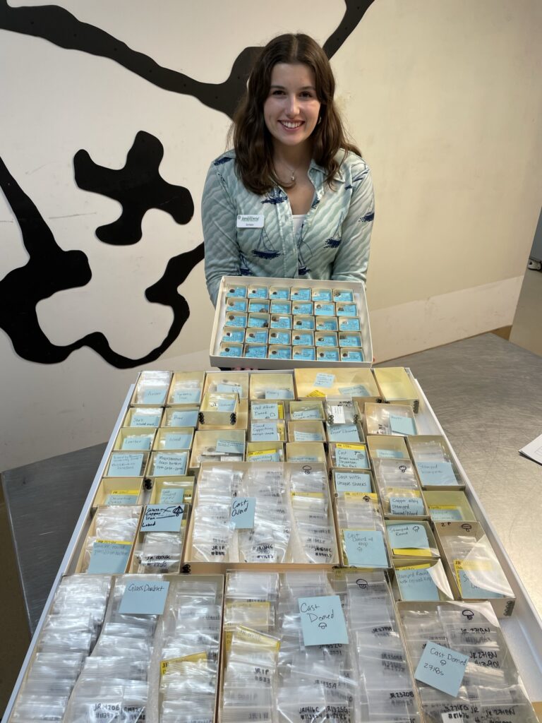 A young, brunette woman stands in front of a drawer full of small artifact boxes with buttons in them