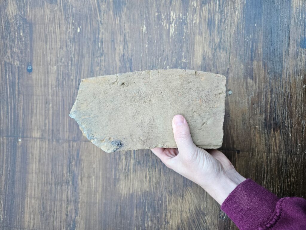 A person holds an irregularly shaped brick above a wooden table. The brick is very rounded on the long side, forming a slight "C" shape.