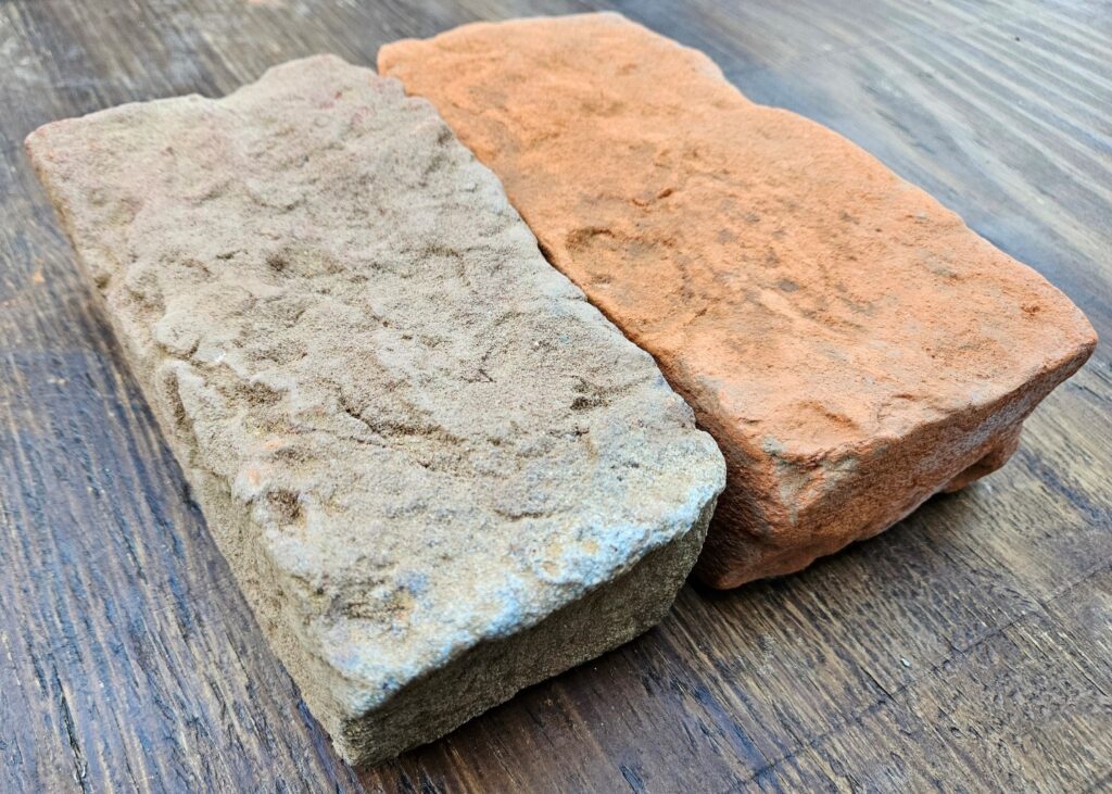 Two bricks are laying next to each other on a wooden table. The brick on the left is tan with a very rough surface. The brick on the right is orange with a smoother surface.
