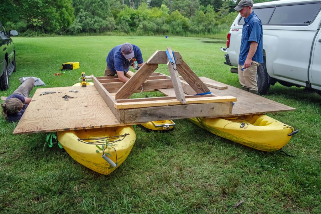 Two yellow kayaks rest on the grass. A plywood platform rests on the kayaks with a triangular structure built on top. Two people are working on the structure.