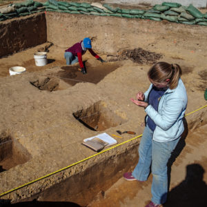 archaeologist examining a find while another archaeologist excavates in the background