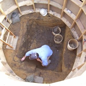 Archaeologist troweling in a square-shaped well surrounded by a circular supporting wooden structure