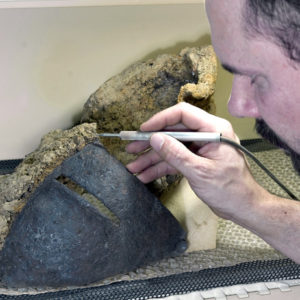 Conservator using an air abrasion tool to remove corrosion on a half-conserved helmet