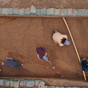 The archaeology team inside two of the squares in the North Church site. The 1608 palisade extension's post molds can be seen running left to right near the bottom of the photo. An early twentieth-century utility line can be seen at the top left and a modern yellow conduit can be seen in the right side of the photo. At top right, Archaeological Field Technician Katie Griffith points to a burial.