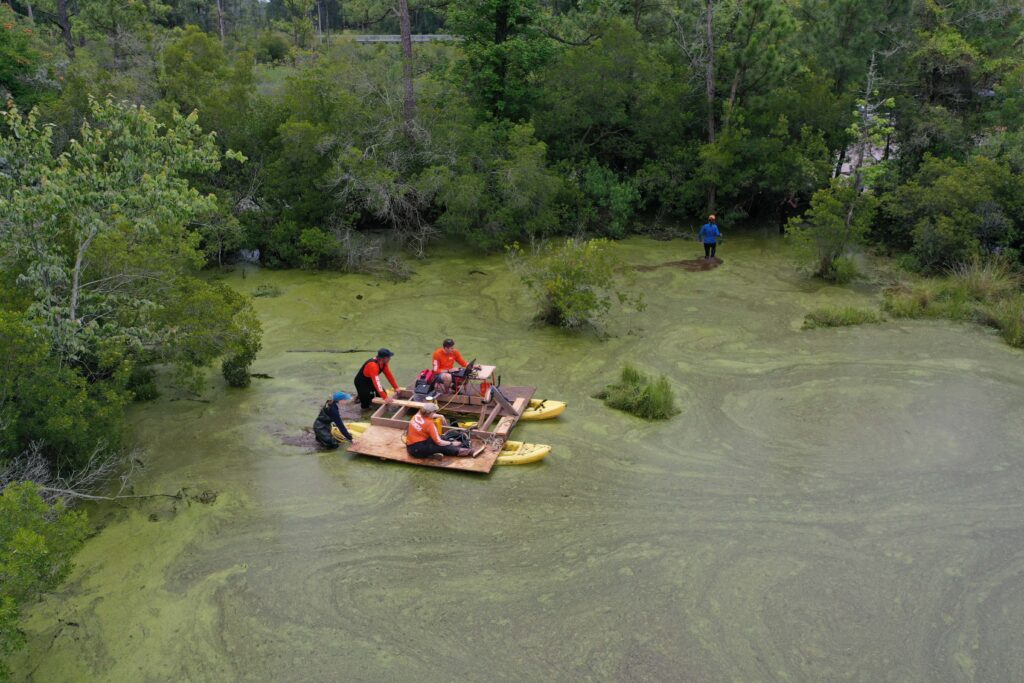 A photo of a group of people in waders pushing the kayak platform through the swampy waters. They're surrounded by brush and trees. 