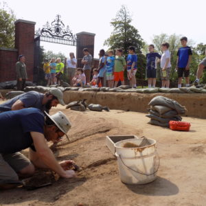 Archaeologists brushing finds in a unit while another gives a tour to school children in the background