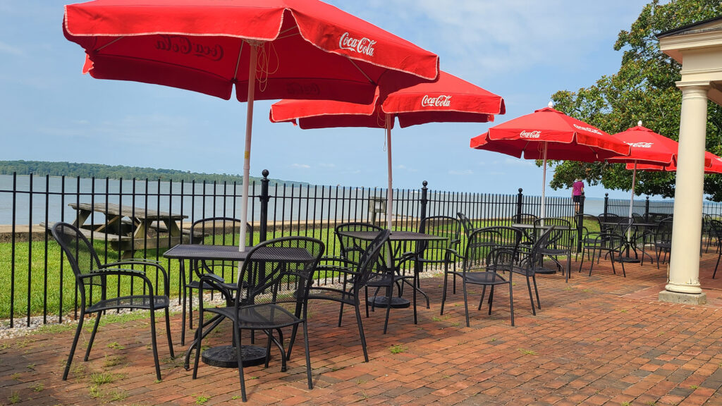 Tables and chairs under umbrellas on the patio of the Dale House Cafe with the James River in the background