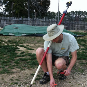 man crouching and hammering nail into the ground while holding a stadia rod