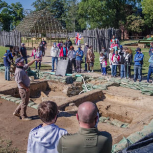 archaeologist standing in an excavation unit and speaking to a group of visitors