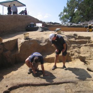 Archaeologist using a trowel to excavate a cellar floor while another observes