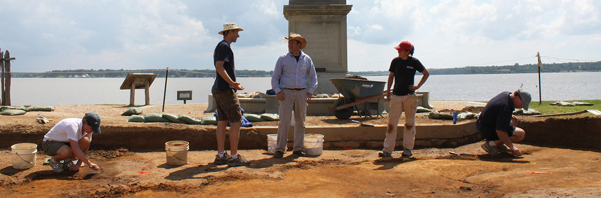 Three archaeologists stand in excavations in front of statue