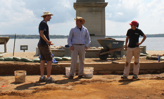 Three archaeologists stand in excavations in front of statue