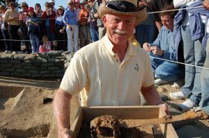 Archaeologist showing an excavated broadsword in a wooden tray to the camera while a group of visitors take photographs in the background