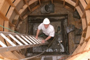 Archaeologist smiling at camera while standing at the base of a ladder inside an excavated well surrounded by a circular wooden support structure