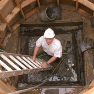 Archaeologist smiling at camera while standing at the base of a ladder inside an excavated well surrounded by a circular wooden support structure